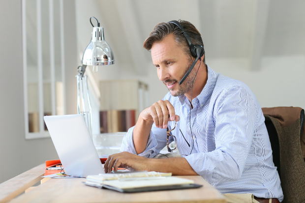 Man in blue shirt on a laptop with a headset on working from home.